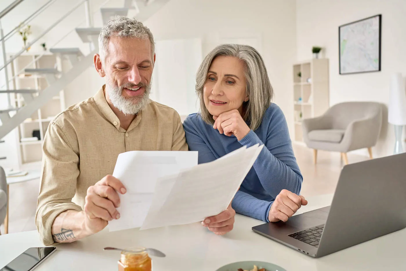 An elderly couple are looking at documents at the kitchen table with their laptop open.