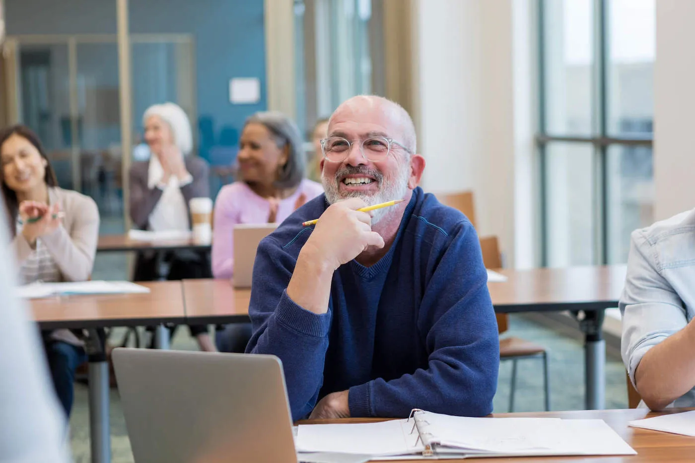 An elderly man wearing a blue sweater smiles in the classroom.