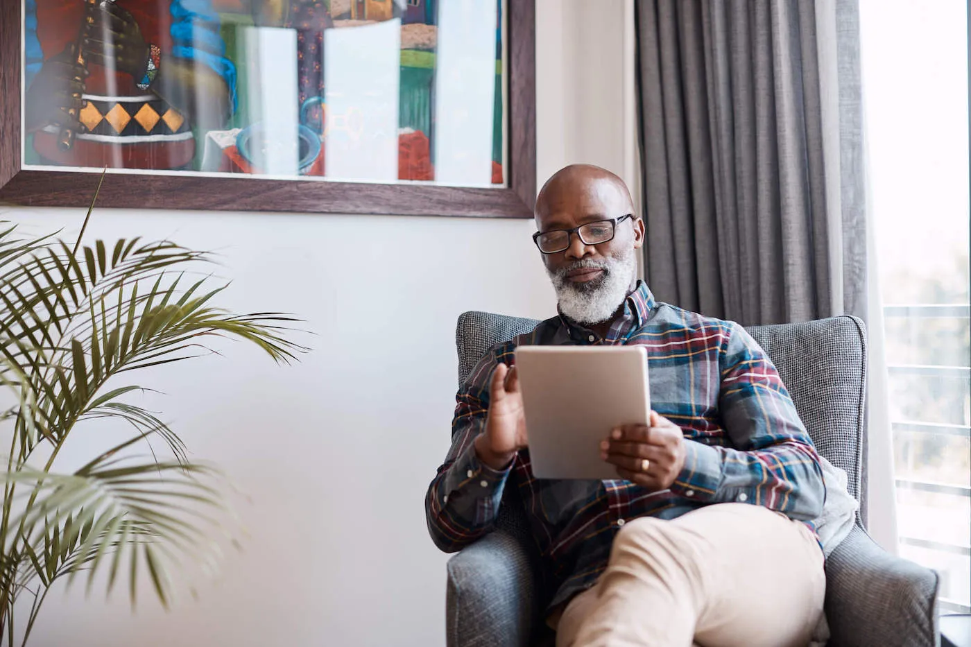 An elderly man uses his tablet while sitting on a chair at home.