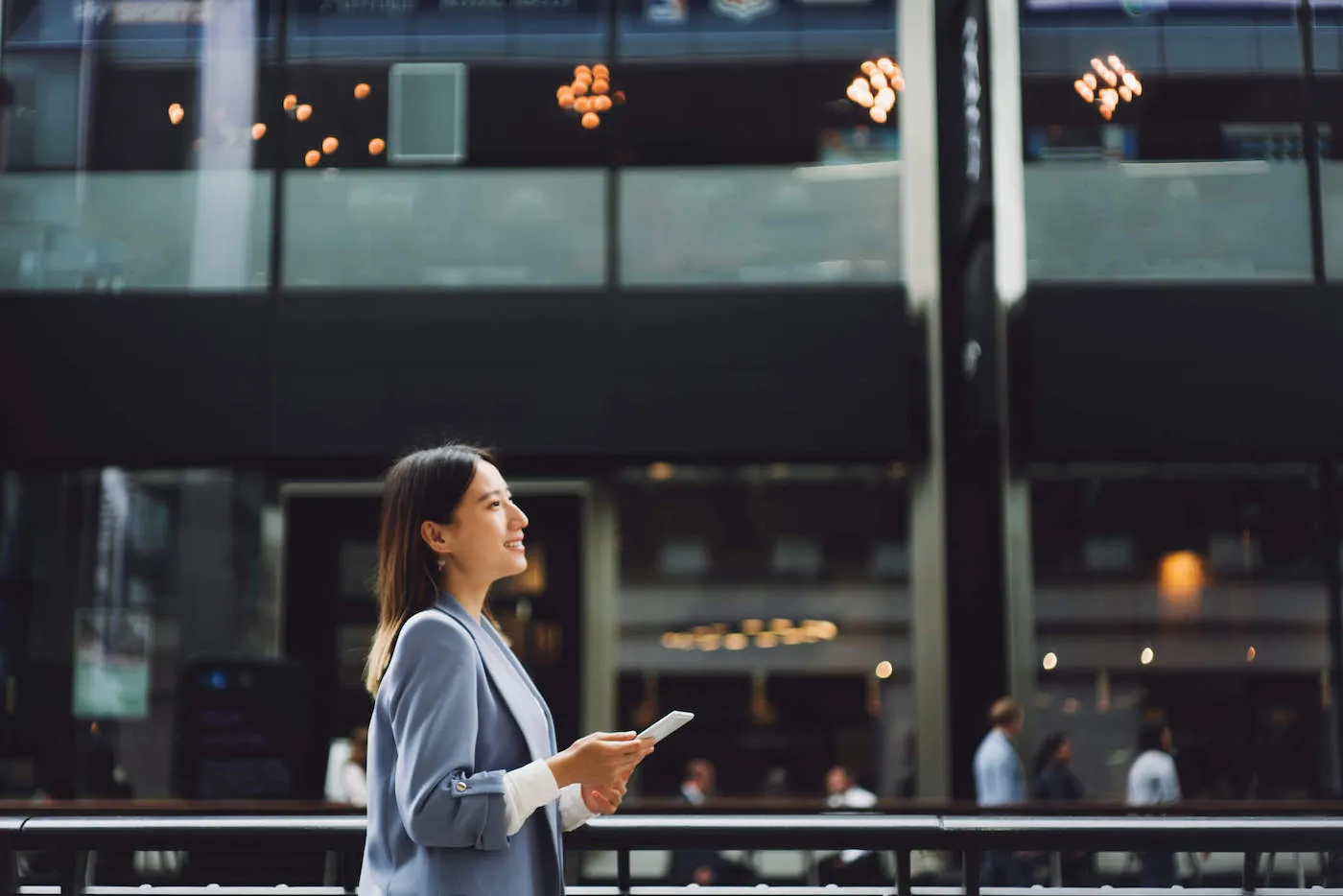 Asian businesswoman walking in a financial area looking up to the sky while holding a smartphone.