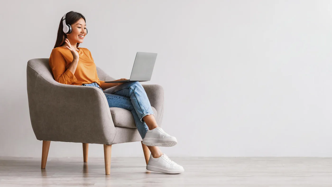 Asian female in headphones using laptop while sitting in armchair.