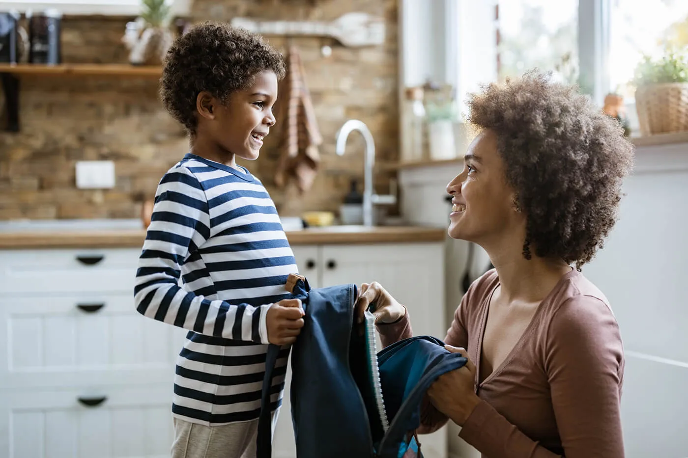 Happy mother talking to her small son while packing his books into a backpack.