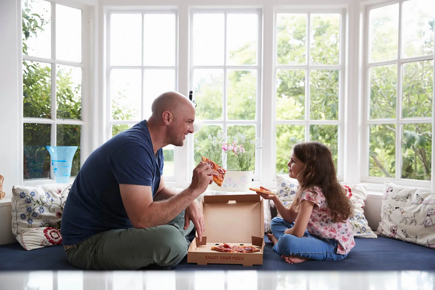 Dad and daughter sit on window seat at home sharing a pizza