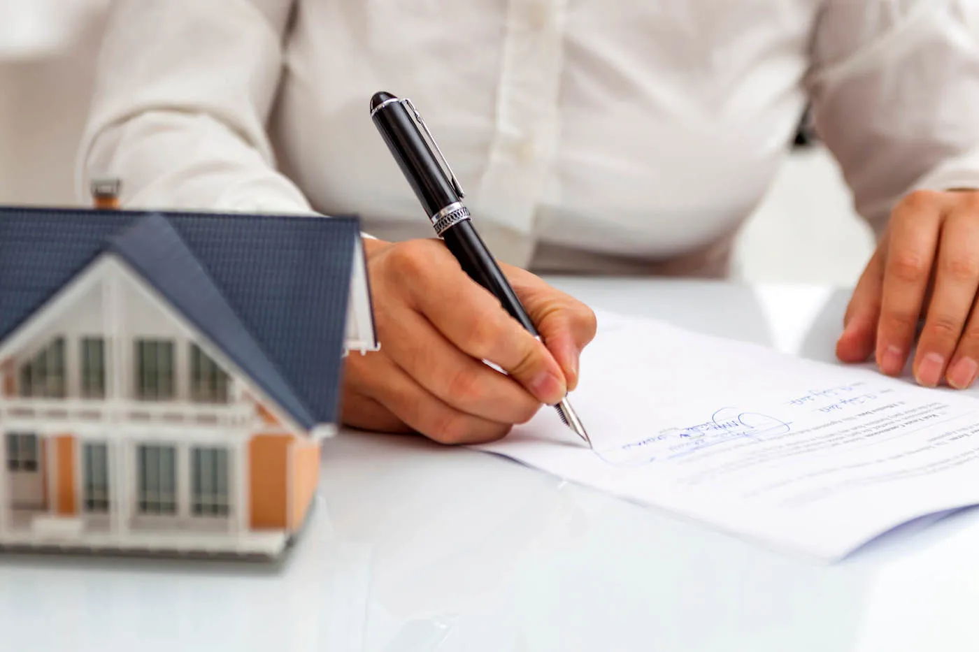 Closeup of a man's hands that is signing a document with a pen next to a miniature house model.