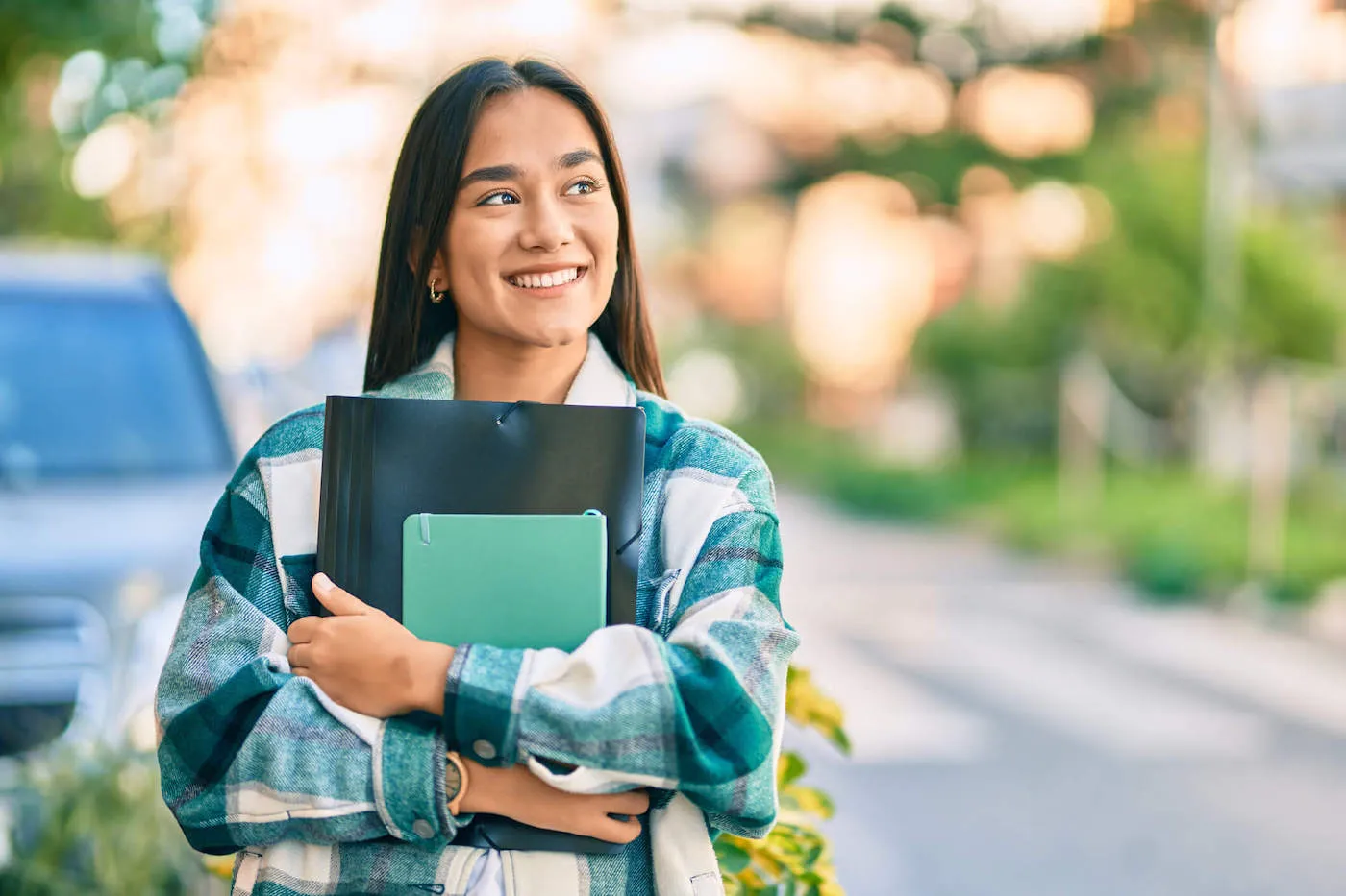 A college student is wearing a striped shirt outside as she smiles while holding her school folder.