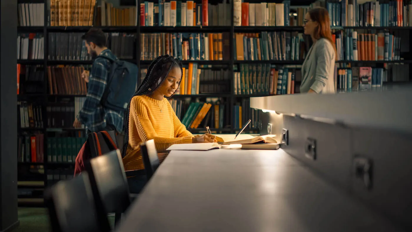 A college student wearing a yellow shirt sits down and studies in the library.