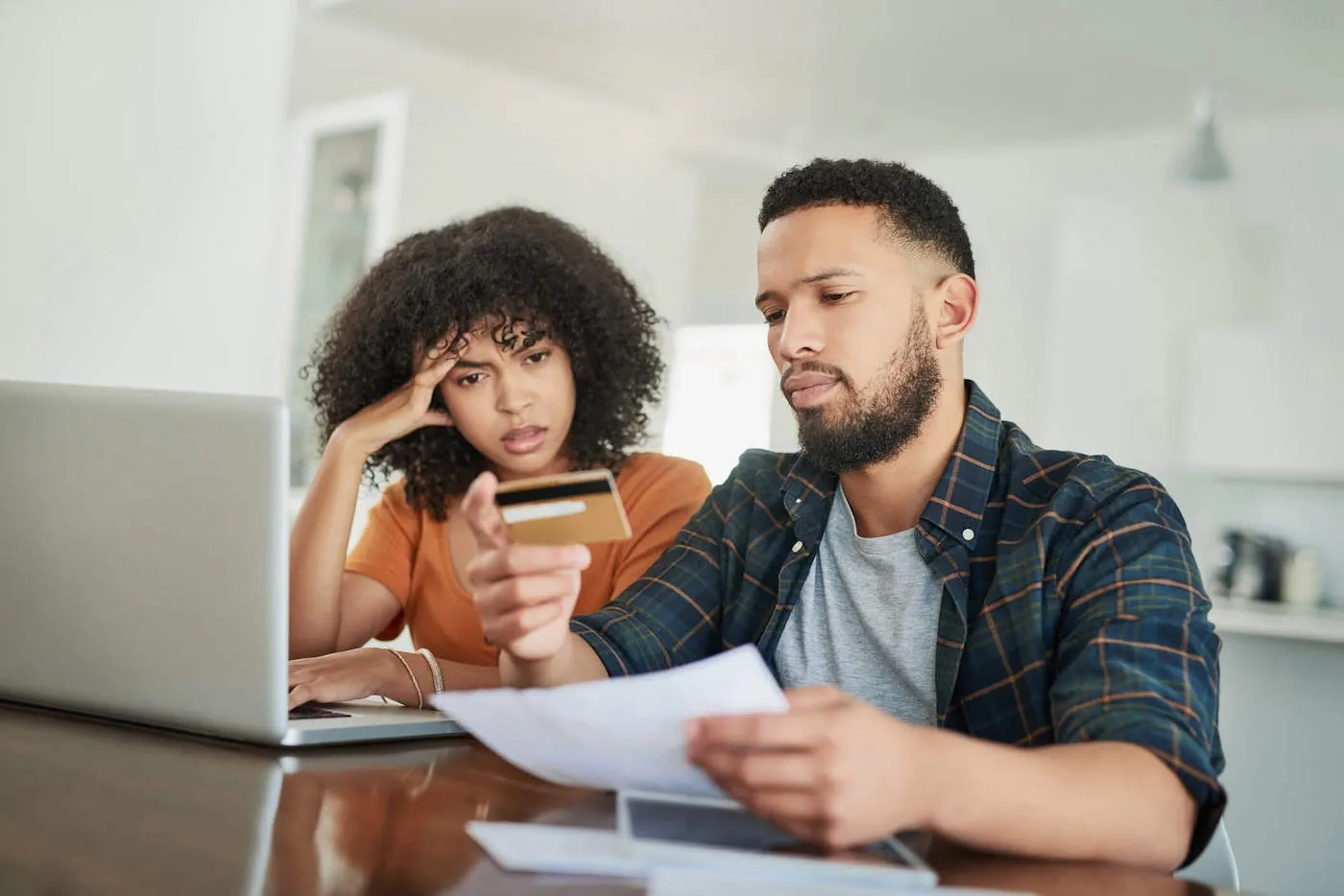 A couple look at a credit card together and frown while documents and a laptop are on the table.