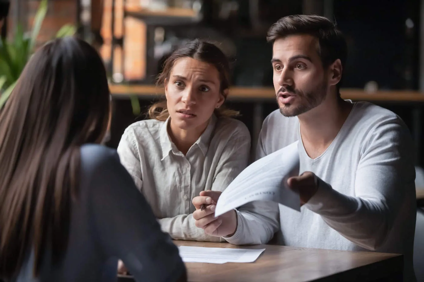 A couple with a surprised look hold documents while talking with a woman from across the table.