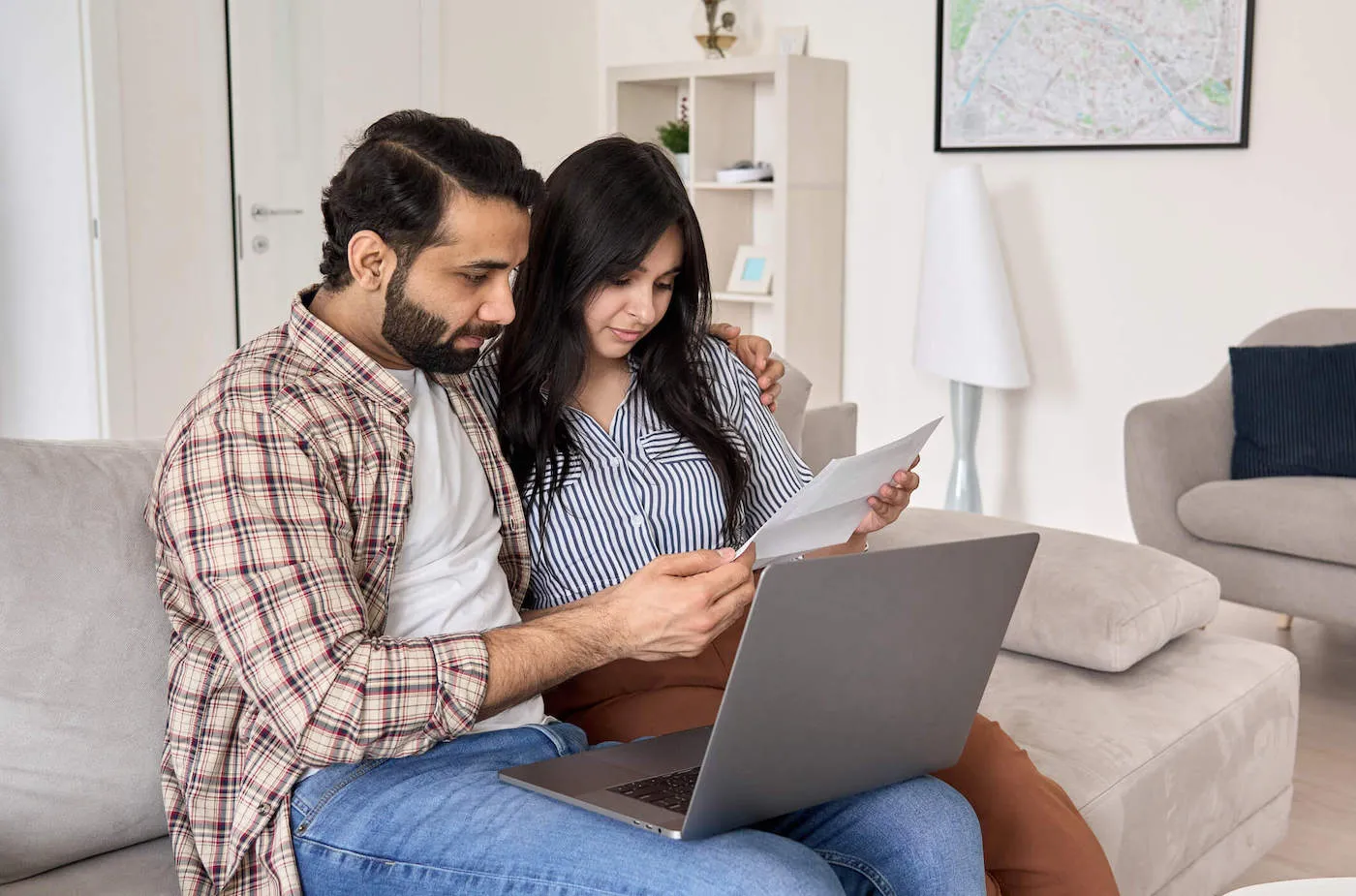 A couple sits on the couch together while looking at a document with their laptop right next to them.