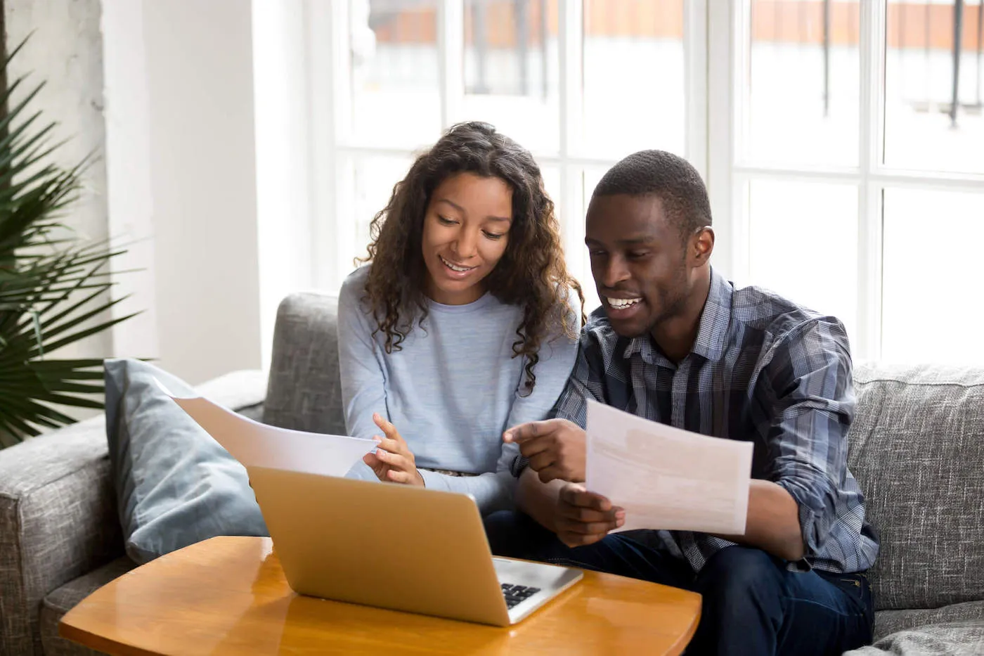 A couple sitting on the couch together smile at a laptop while holding documents.