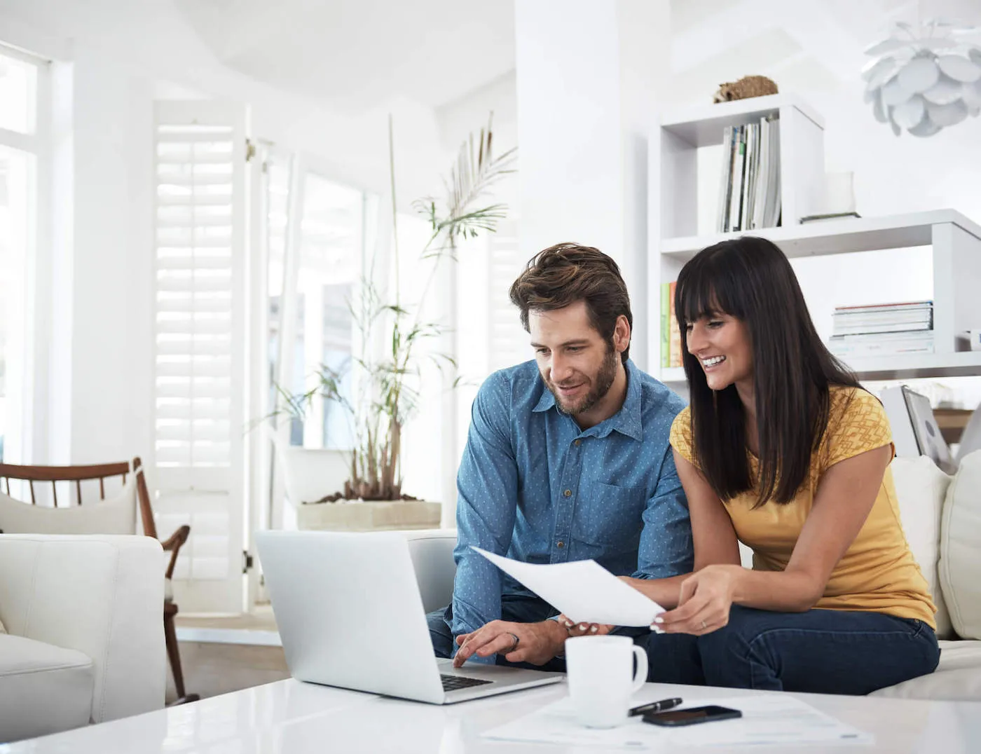 A couple smile together at a laptop while sitting on the couch.