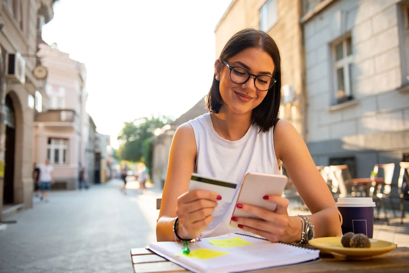 A woman smiles as she uses her phone and holds her credit card above her notebook while having breakfast outside.