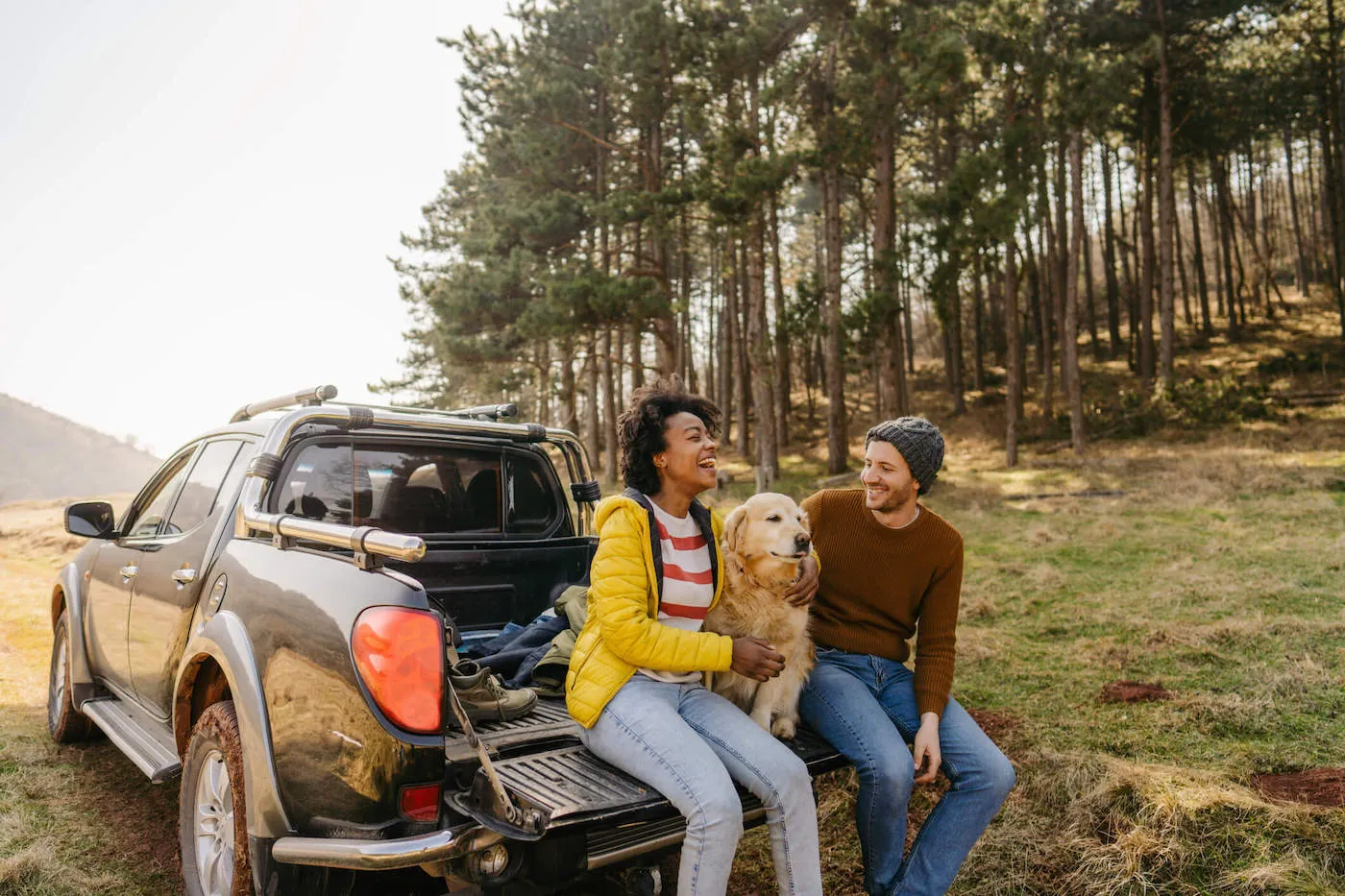 man and woman sitting in the back of a pickup truck with a dog among the woods