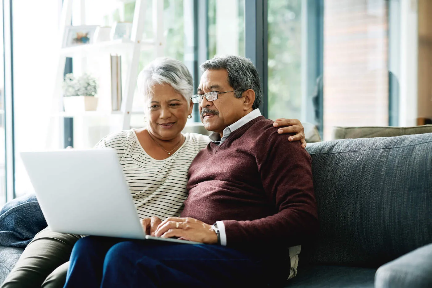 An elderly couple sit on the couch together while they both look at a laptop screen.