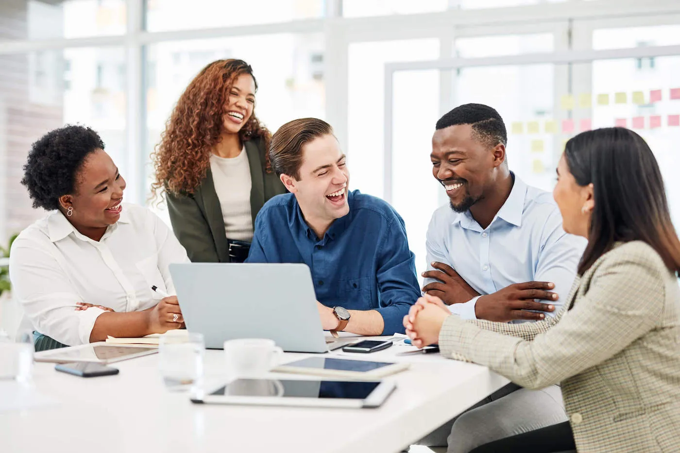 A group of five employees are huddled around a table and laughing with each other at the office.