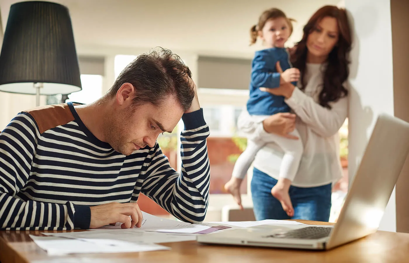 A stressed man sitting at a table looking at papers and his laptop while a woman holding a baby looks at him with sympathy.