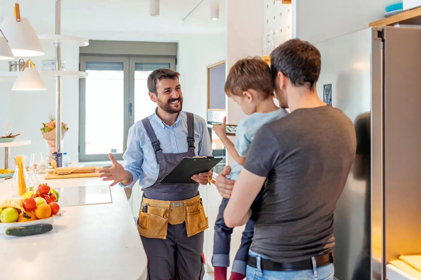 A home inspector hold a clipboard while talking to a father carrying his son in the kitchen.