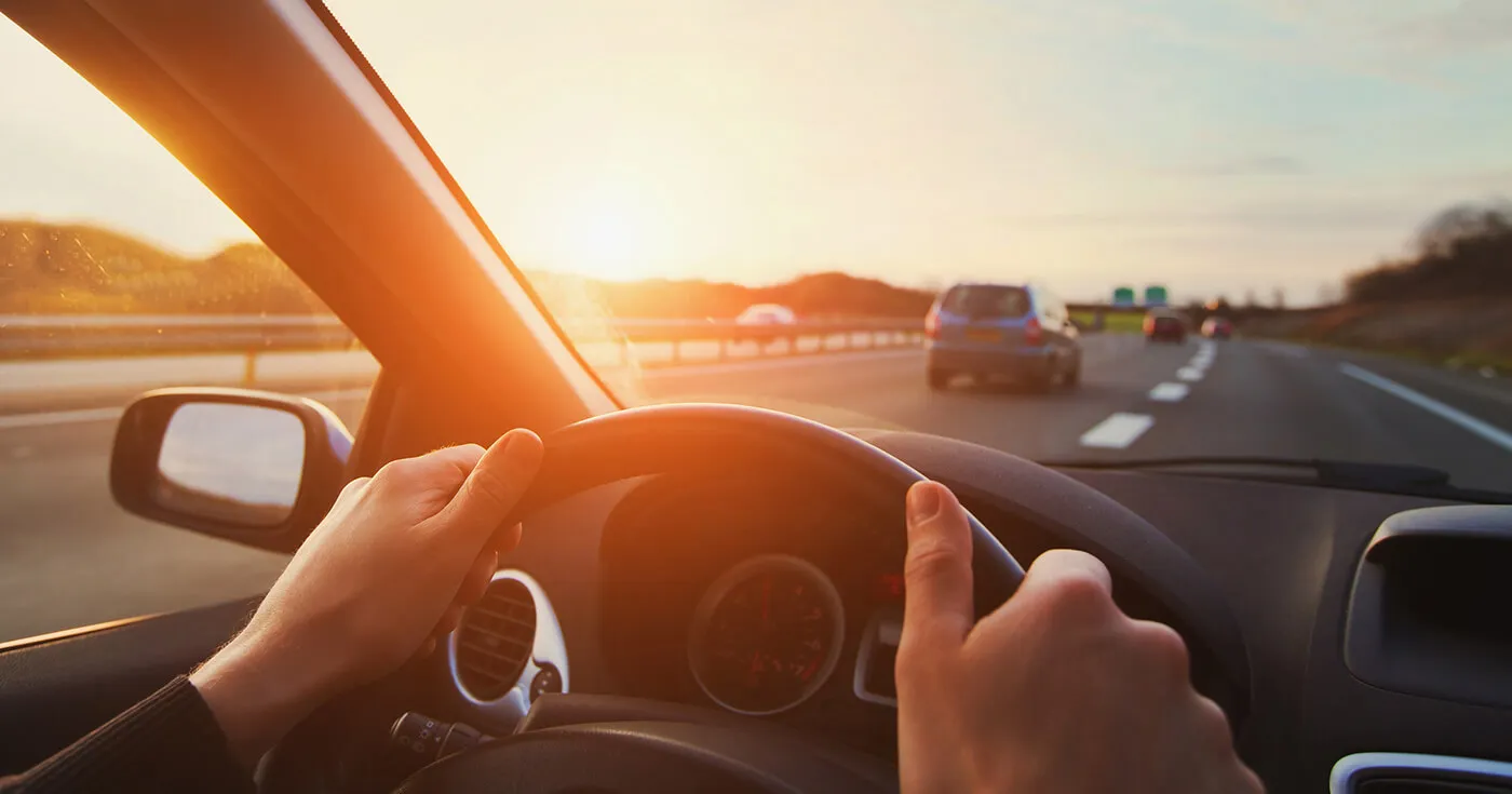 Shot of a driver's hands on a steering wheel, driving on a road at sunset.