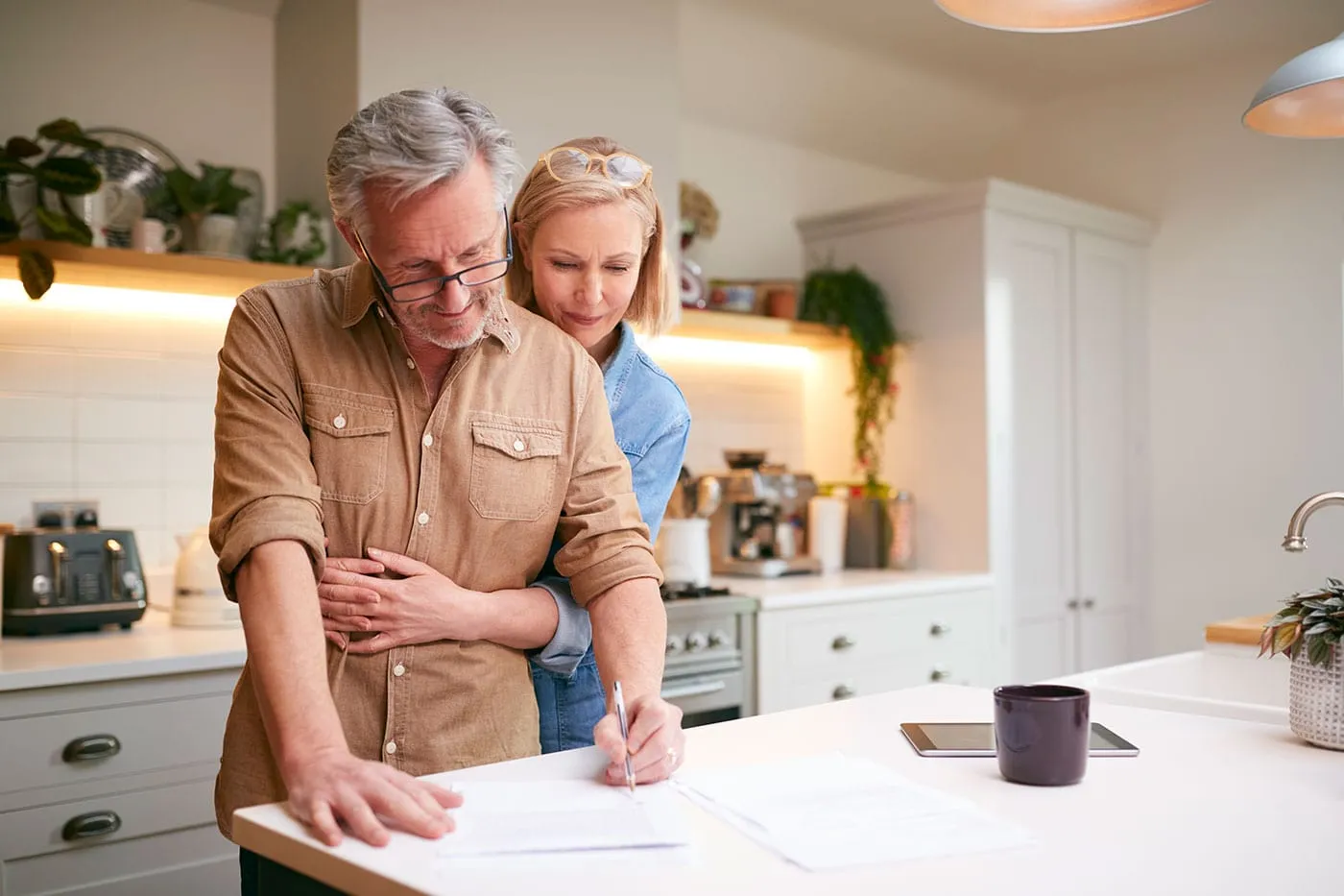 An elderly couple is at their kitchen signing a document while the woman embraces the man.