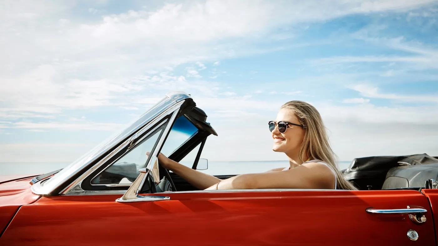 A happy young woman wearing sunglasses in a red convertible driving by the ocean, enjoying a summer’s road trip.