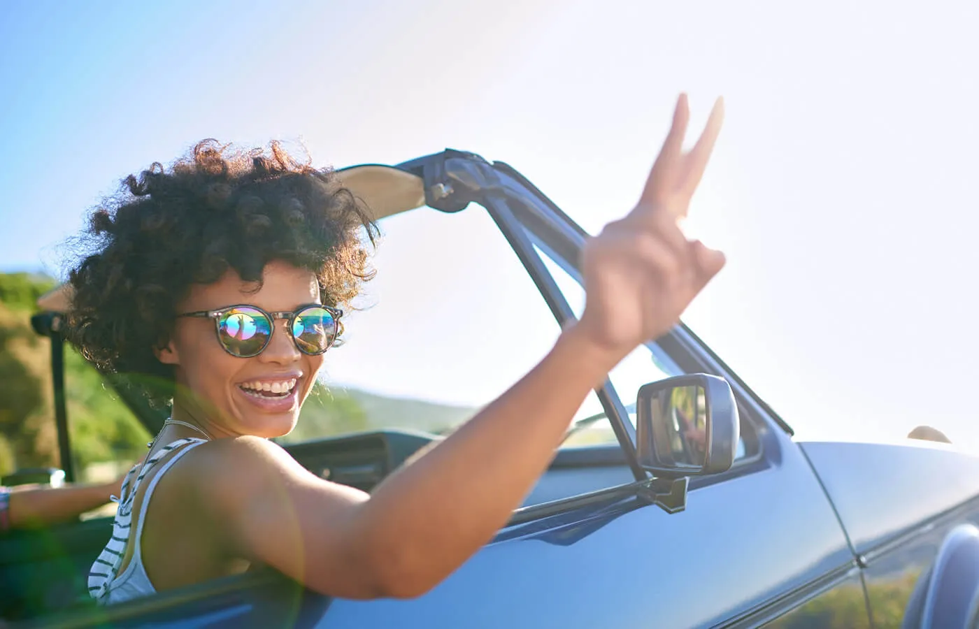 smiling woman in sunglasses holding up two fingers while riding in a convertible