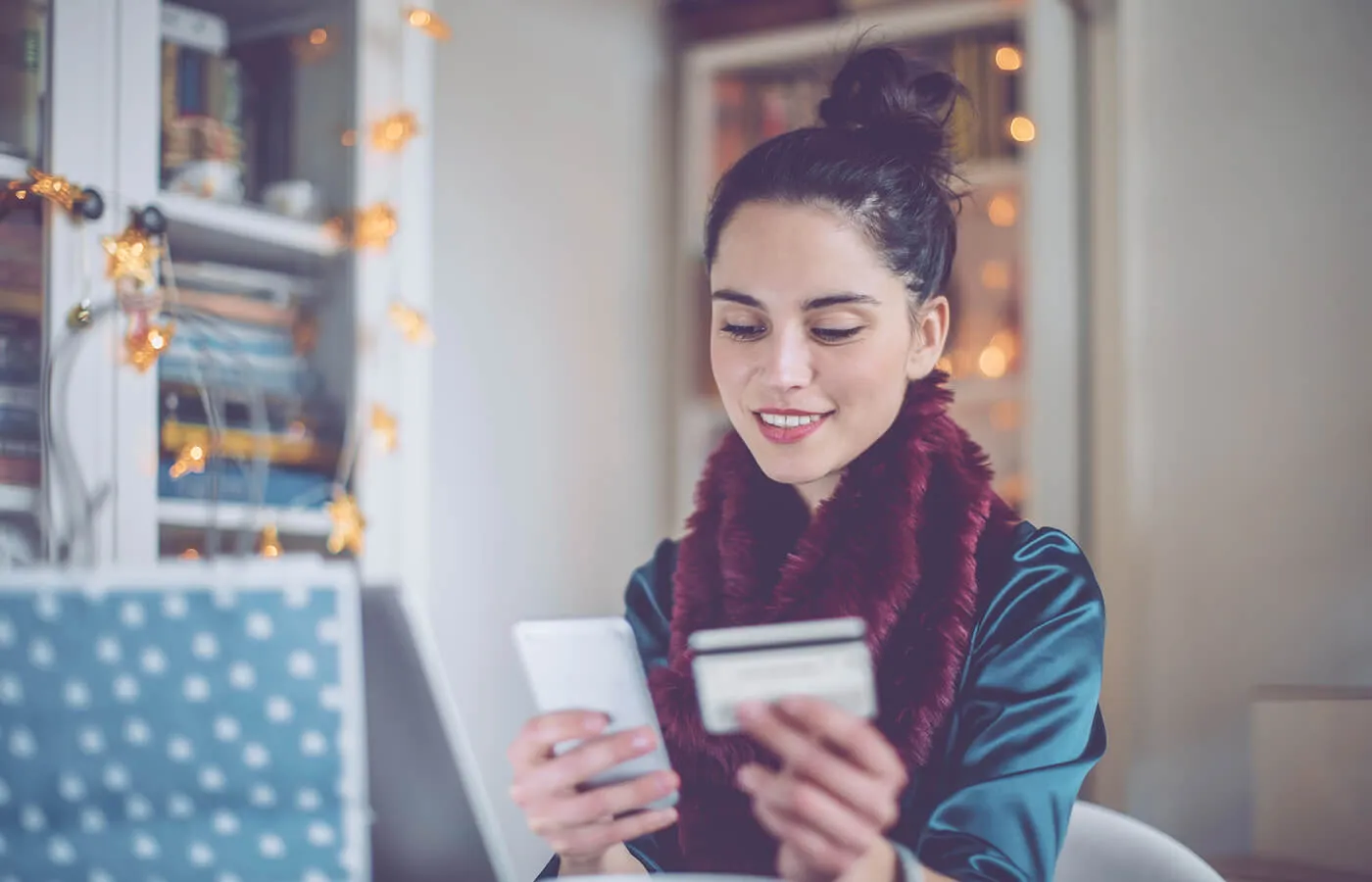 A woman wearing a blue shirt and red scarf smiles as she holds her phone and credit card.