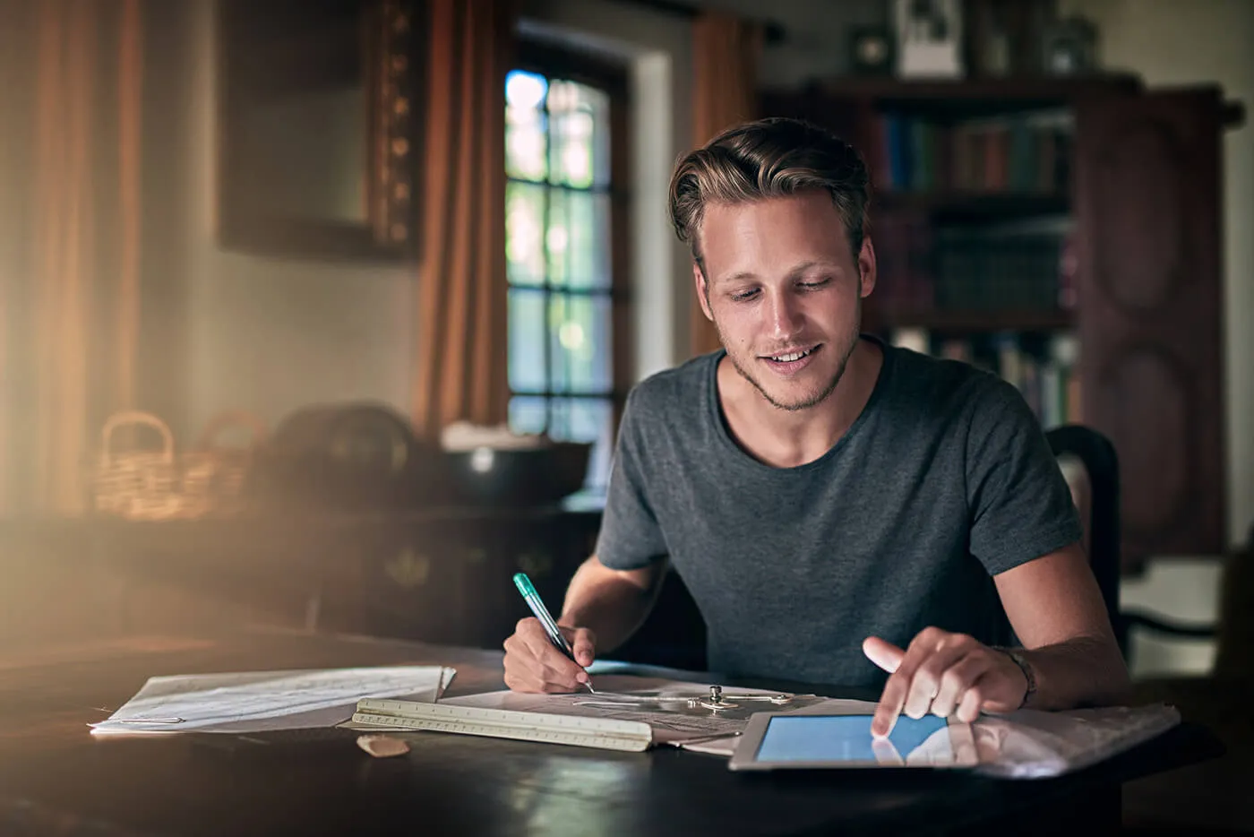 A young man smiles while looking at his tablet and writing on his notebook.