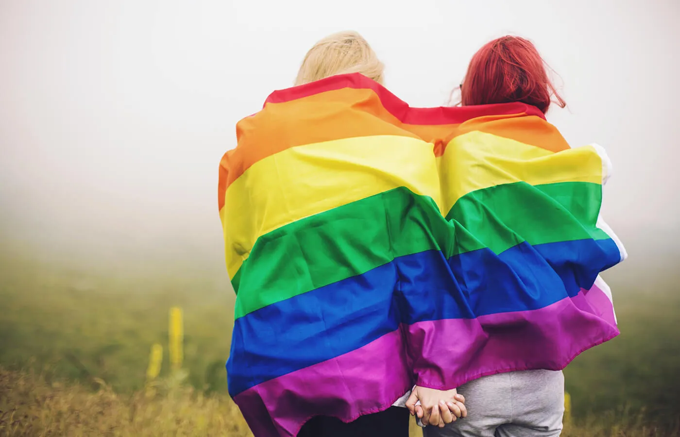 A queer couple wrapped in a pride flag sitting on a misty mountaintop.