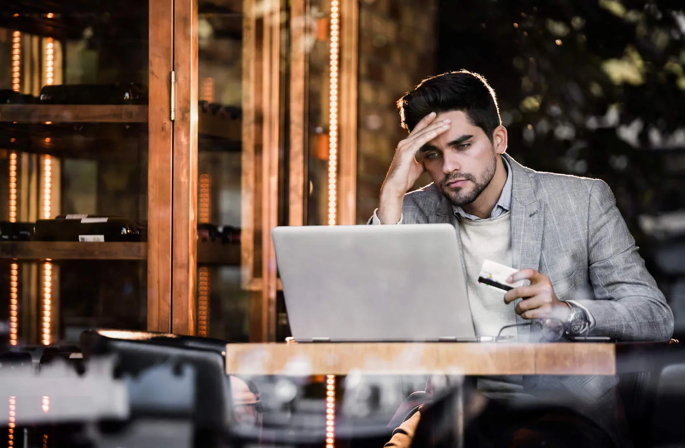 A man wearing a gray suit frowns while looking at his laptop and holding his credit card.