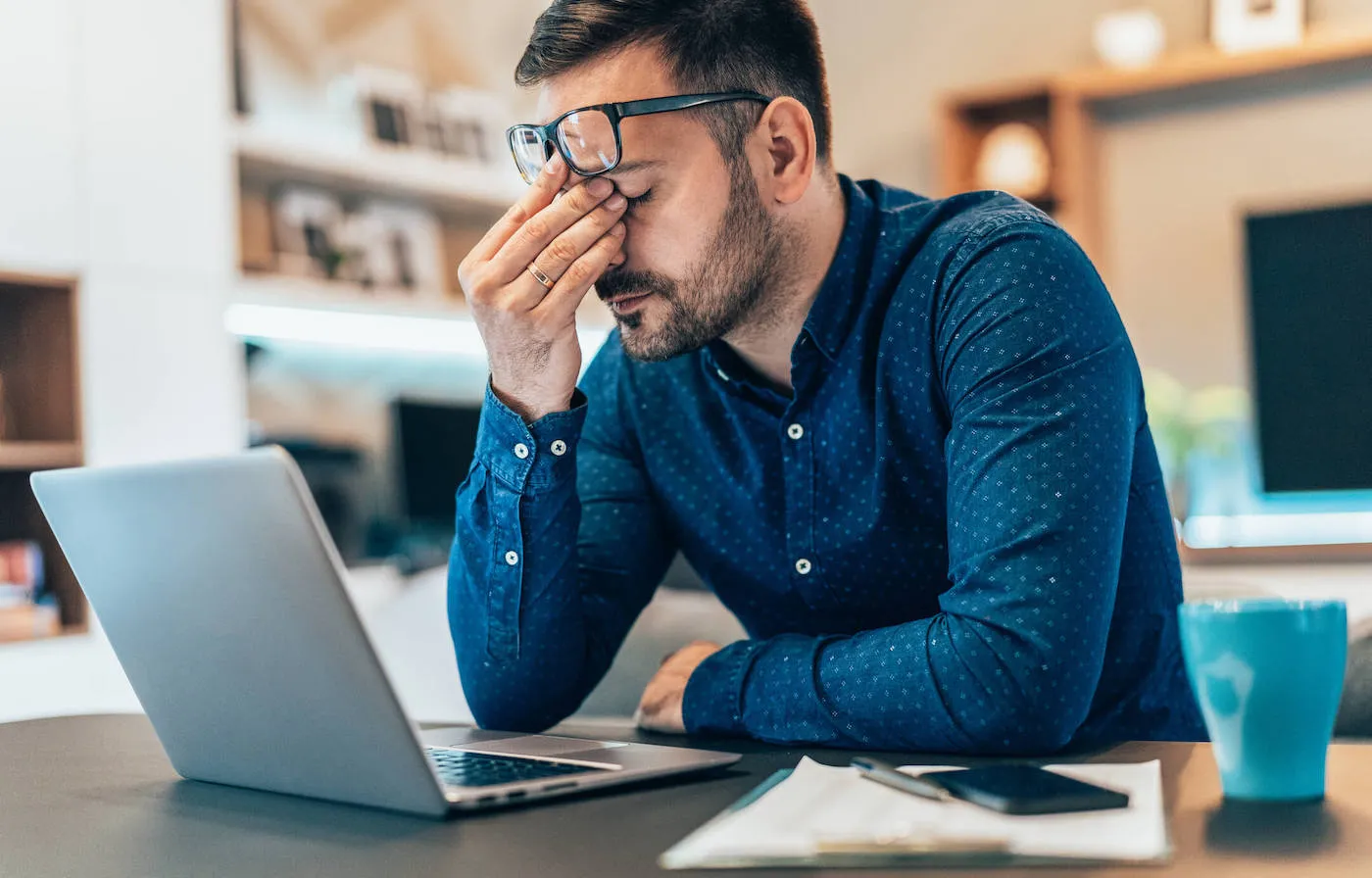 A man wearing a blue shirt has his hand to his face with his laptop open in front of him.