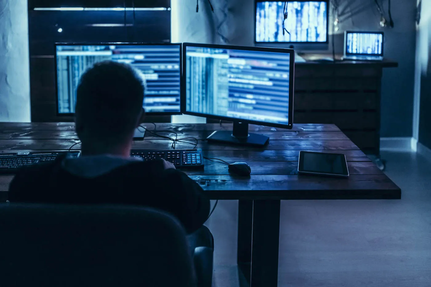 A man sitting in a dark room looks at his desktop computer screens.