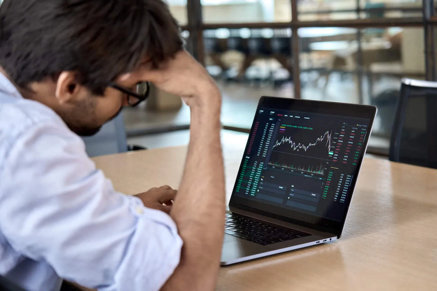 A man wearing glasses leans on his hand while looking at a stock chart on a computer.