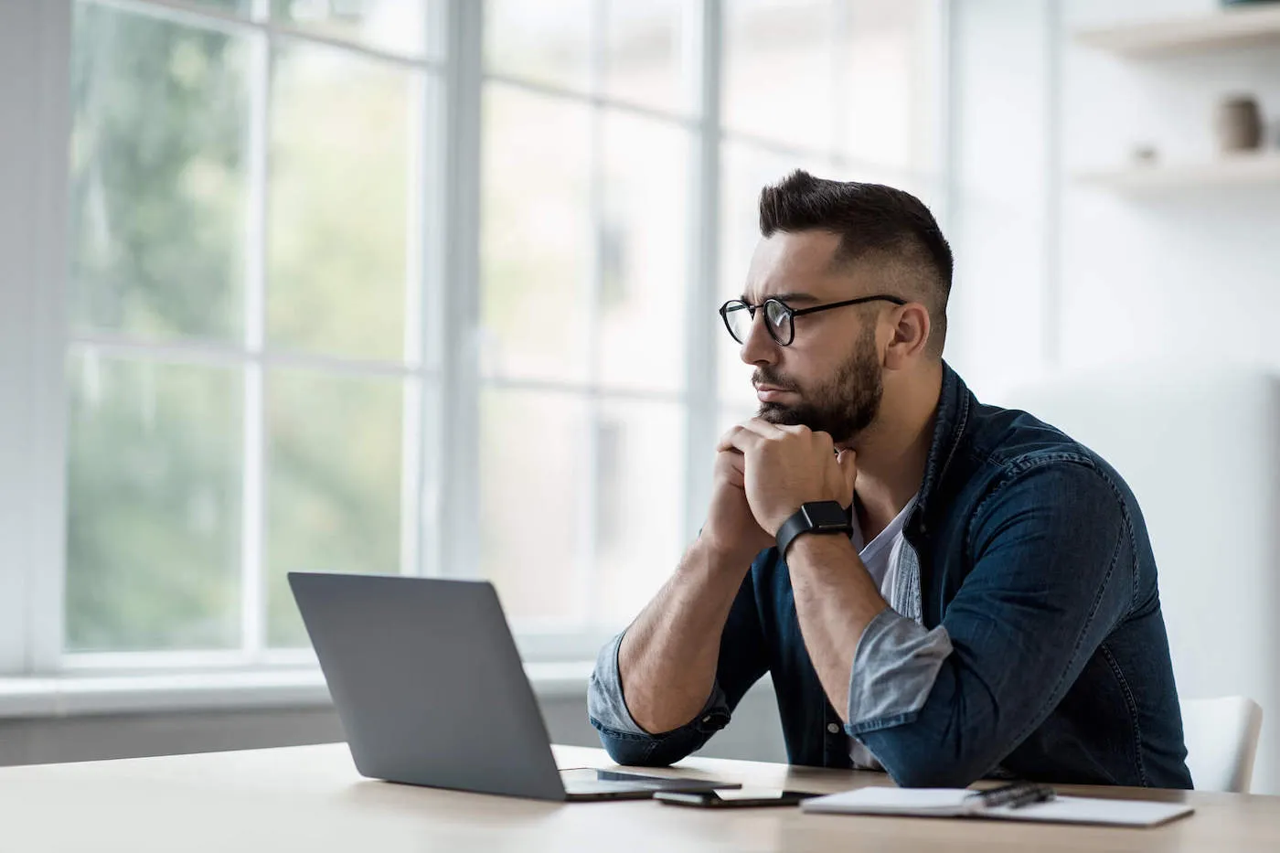 A man wearing a blue shirt and black glasses has a concerned look on his face as he stares out the window with his laptop open.