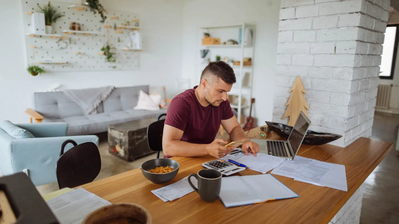 A man wearing a red shirt looks at a document on the kitchen table while his laptop is next to it.