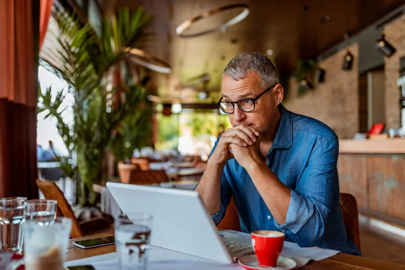 A man wearing a blue shirt is looking out the window of a coffee shop with his hands to his mouth as he has his laptop open.