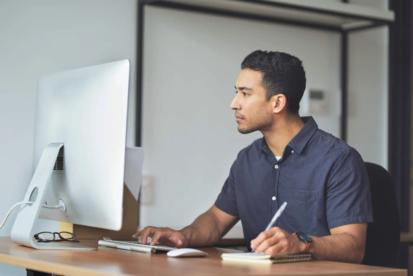 A man wearing a blue shirt looks at his computer screen while writing something down in his journal.