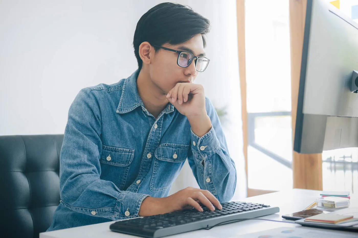 A man wearing a blue denim shirt and glasses looks at his computer screen.