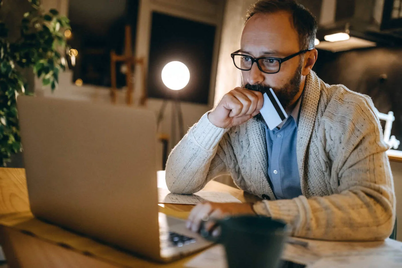 A man wearing glasses looks at his laptop while holding a credit card.