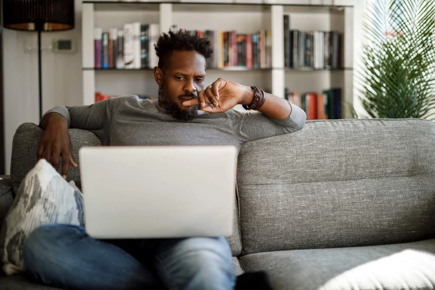 A man wearing a gray shirt sits on the couch while looking at a computer.