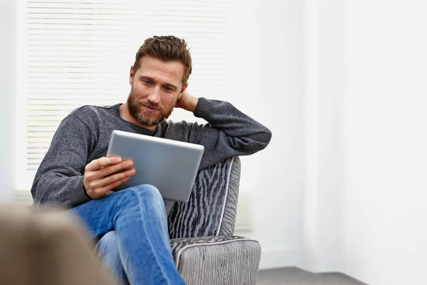 A man wearing a gray sweater sits at a chair while looking at a tablet he's holding.