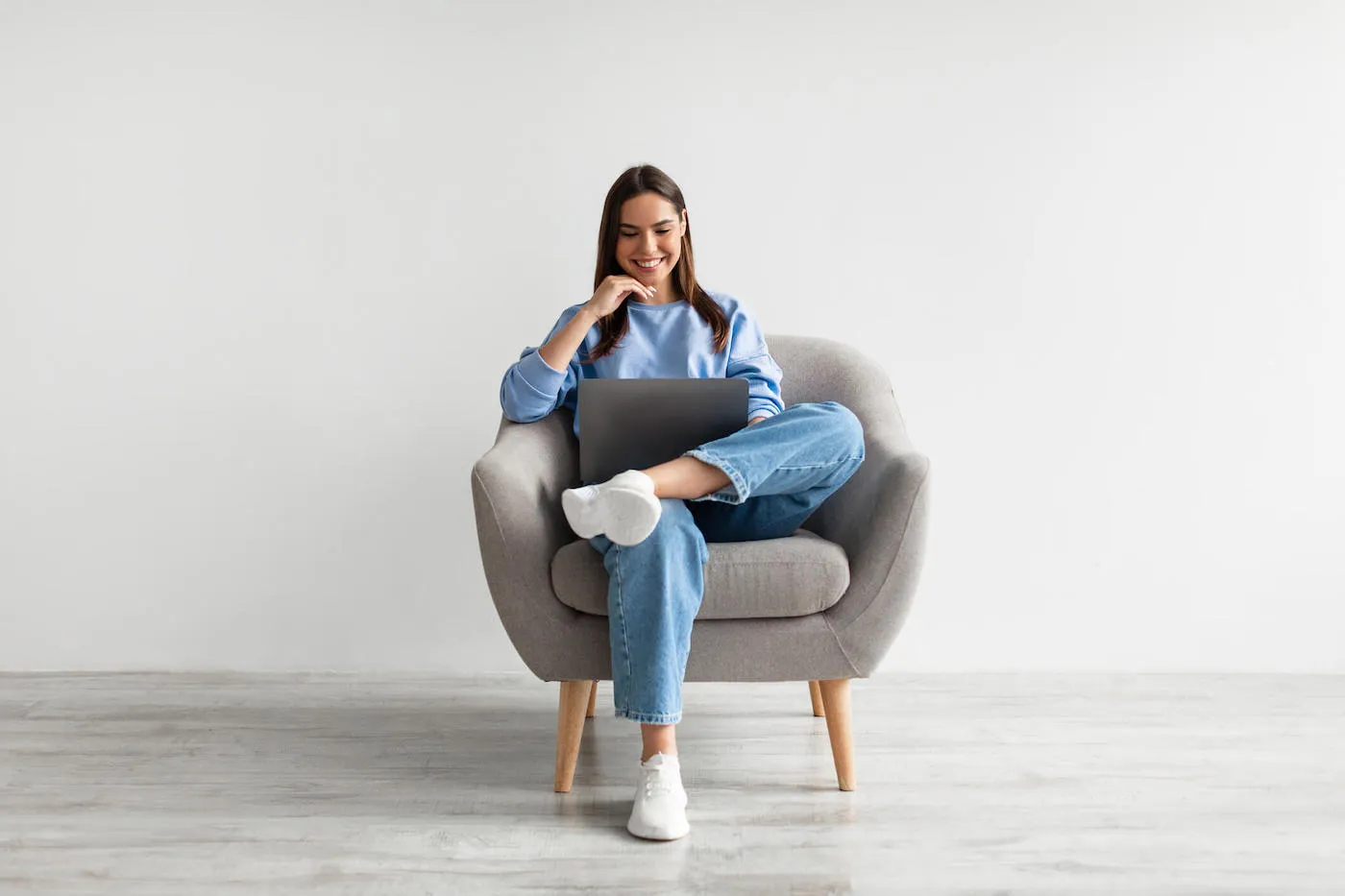 Millennial Caucasian lady in casual wear using laptop, sitting in armchair, having online conference against white wall.