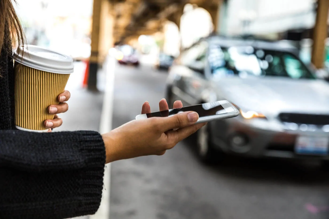 A woman checking her phone and holding a coffee outside next to the street.