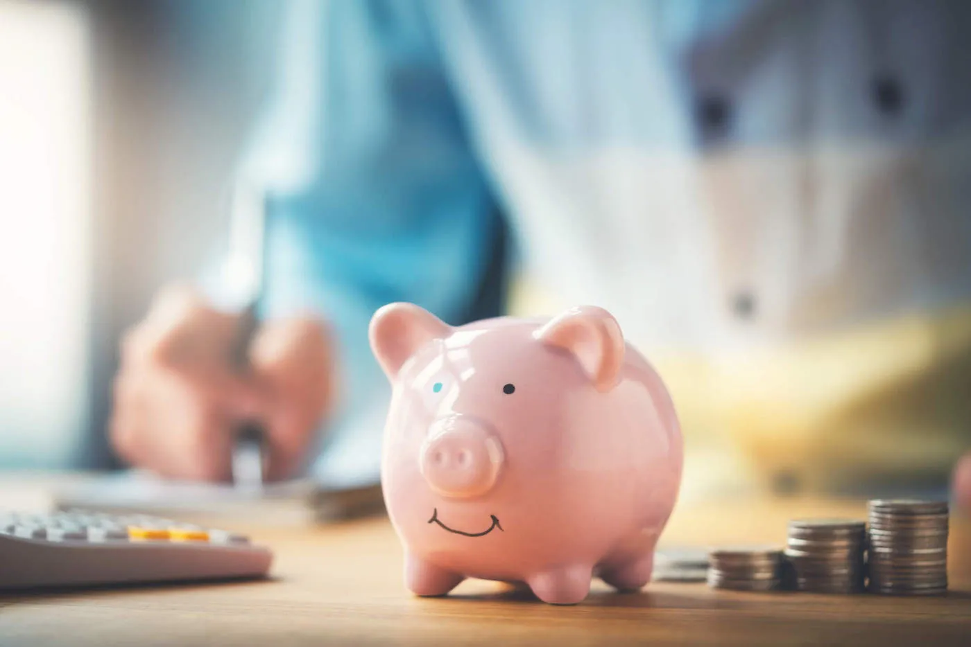 A pink piggy bank sits on the table next to a calculator and coins with a person taking notes in the background.