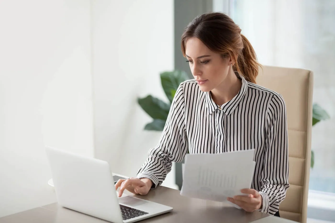 Serious focused businesswoman typing on laptop holding papers preparing report.