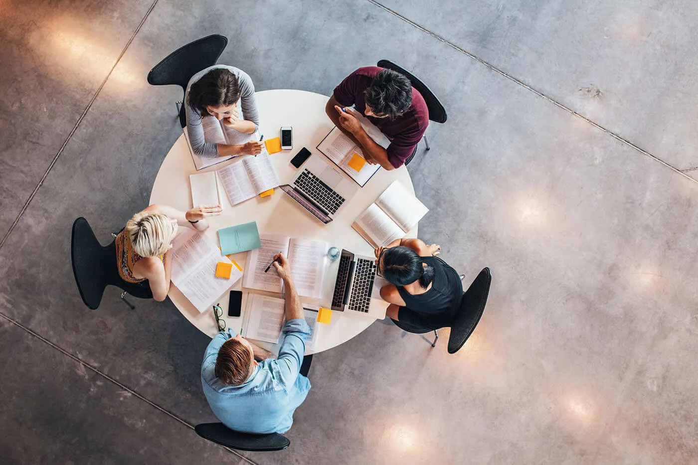 A group of students gather around a table together while having their laptops and documents on the table.