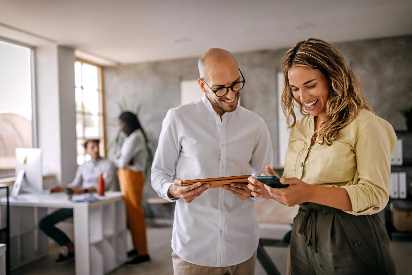 Two coworkers are smiling and looking at a phone while two of their other coworkers are discussing in the background.