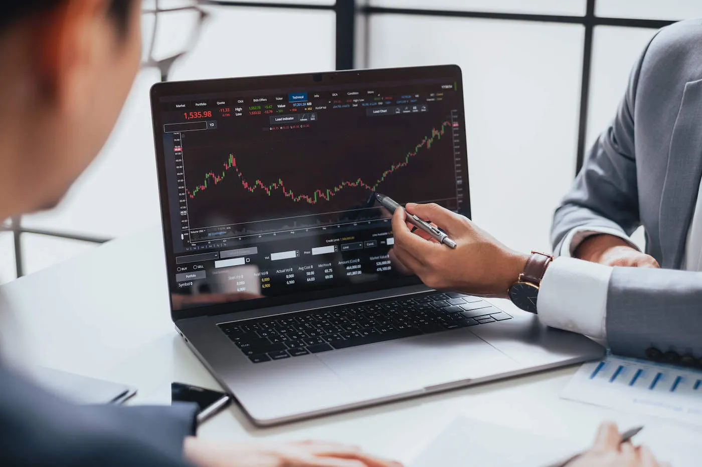 Two people wearing suits are looking at an investment graph on a laptop screen while one person points at the graph with their pen.