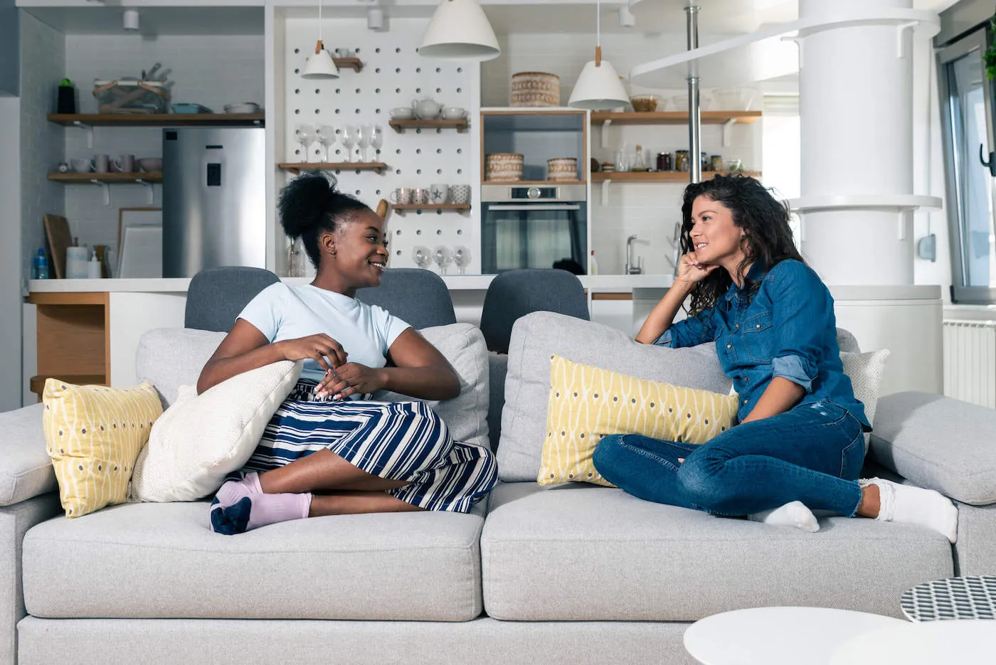 A woman wearing a blue shirt talks to another woman wear a light blue shirt across from her while sitting on a gray couch.