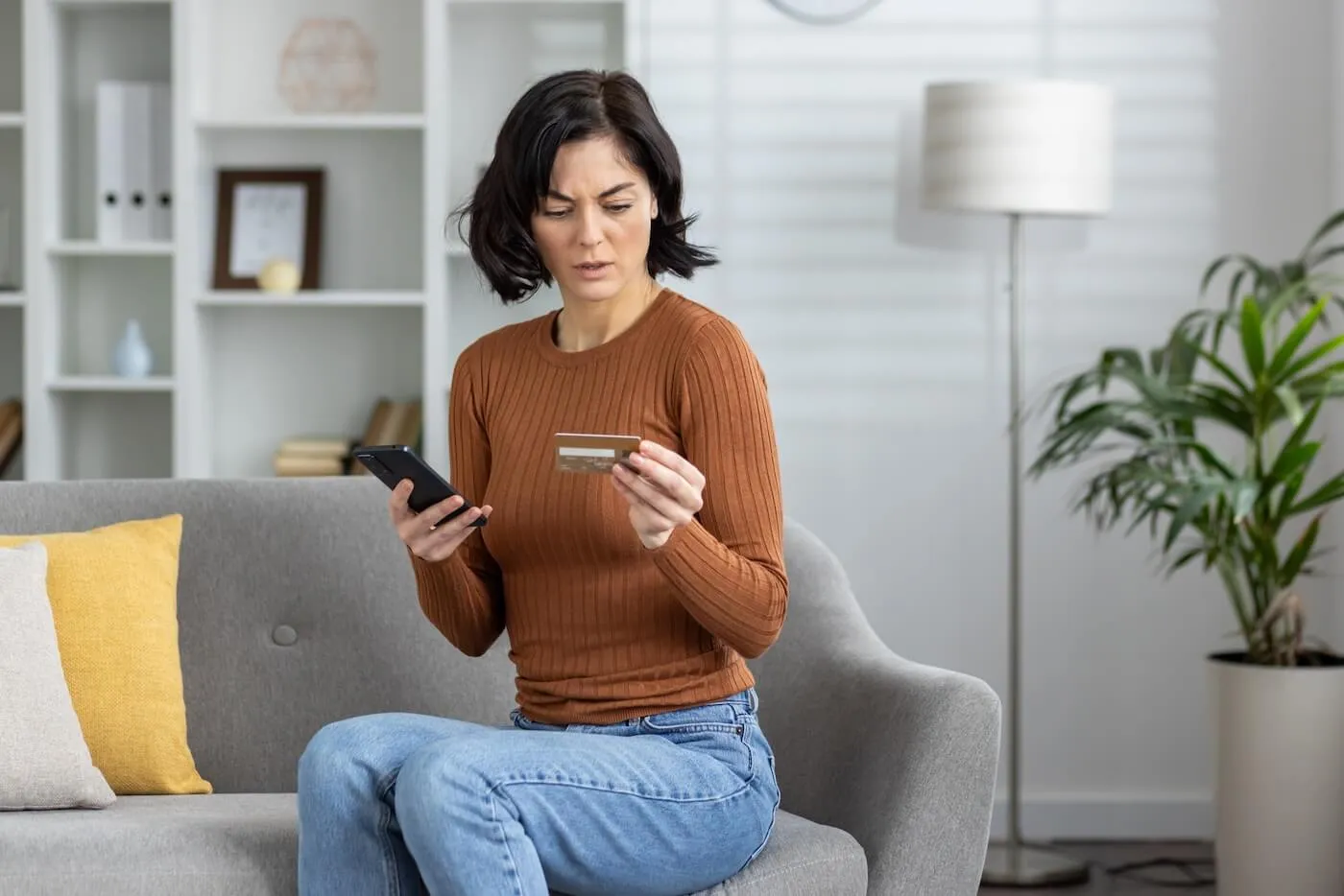 Concerned woman sitting on the sofa and holding a smartphone and a credit card