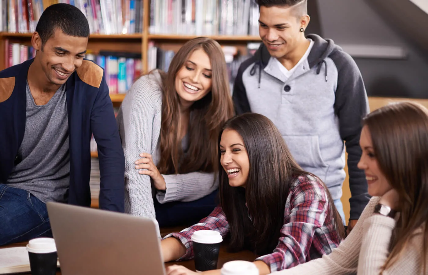 A group of students in a library, smiling and working on a laptop.